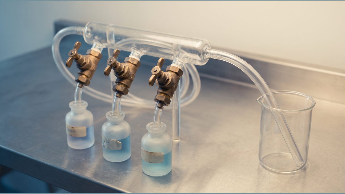 Three brass valves and clear tubes feeding a transparent manifold on a steel bench.